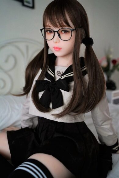 A love doll with glasses and long hair in a sailor outfit sits on a bed, resembling the Milly 150cm Cute Student.