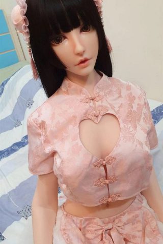 Momo – 165cm Elsa Babe silicone sex doll wearing a pink floral outfit with a heart cutout, seated on a bed.