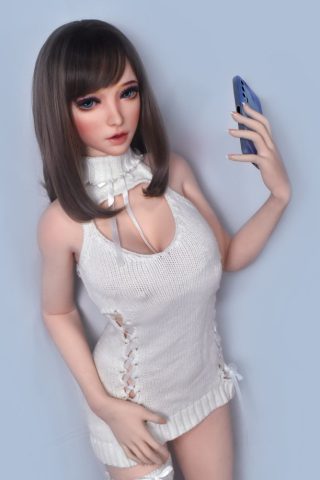Yoshida – 165cm Elsa Babe silicone sex doll wearing a white knitted dress, holding a smartphone, with long brown hair and blue eyes.