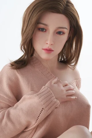 Yvonne F Cup Zelex sex doll, 25, wearing a light pink off-shoulder sweater, with long brown hair and soft facial features.