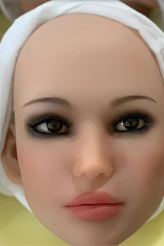 Doll head with a smooth, bald surface, featuring large, dark eyes and soft pink lips, wrapped in a white cloth.