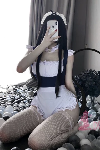 Person in blue maid outfit kneeling on bed with black and white accents, taking selfie in mirror