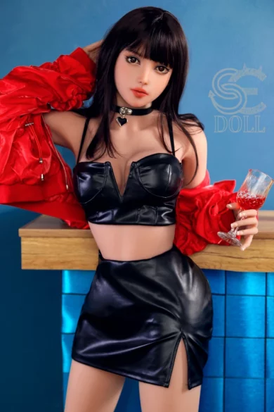 Sex doll wearing a black leather outfit with a red jacket, holding a glass near a wooden counter against a blue backdrop.