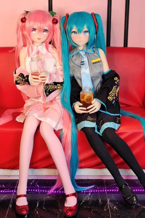 Two anime-style sex dolls with pink and blue hair, B Cup sizes, Heads 74 and 75, sitting on a red sofa holding drinks and smiling.