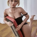 Blonde Gymnast Victoria 150cm B Cup Irontech TPE doll kneeling on floor in black outfit, leaning against red-backed chair with neutral expression