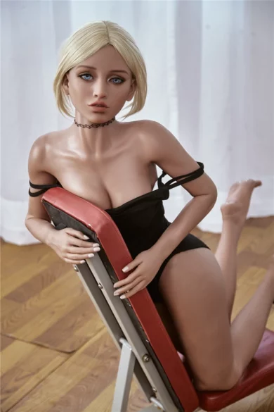 Blonde Gymnast Victoria 150cm B Cup Irontech TPE doll kneeling on floor in black outfit, leaning against red-backed chair with neutral expression