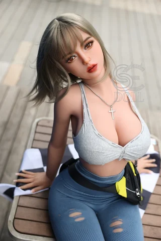 Melody.C 161cm F Cup doll seated on a wooden surface, wearing a gray crop top and blue leggings, with a cross necklace.