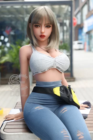 Blonde sex doll wearing gray crop top and blue leggings sits on an outdoor bench.