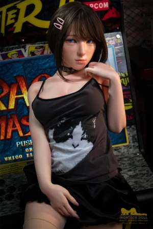 Sex doll with brown hair and blue eyes in black tank top stands before arcade machine