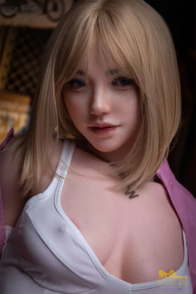 S39 169cm B Cup Irontech love doll with blonde hair in a white top and pink jacket stands in dim lighting