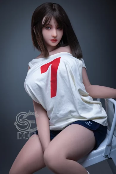 Phoebe C 157cm H Cup #102 SE Doll with dark hair in a white shirt with red number 7 seated on a chair against dim lighting