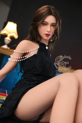 Queena.B 166cm C Cup doll wearing a black dress with pearl accents, seated with a lamp in the background.