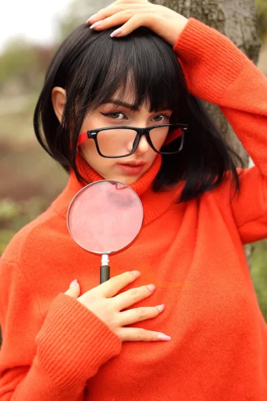 S44 167cm F Cup Tanned sex doll in an orange sweater and glasses, holding a magnifying glass in a playful outdoor setting.