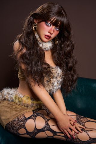 A dark-tanned figure sits on a green couch, wearing a fur-trimmed outfit and fishnet stockings, with long wavy hair and bold makeup.