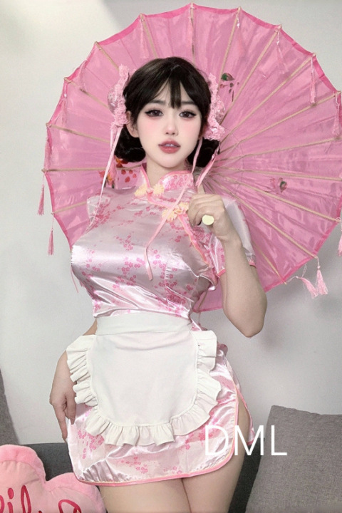 Sex doll in pink floral cheongsam lingerie with white apron, holding pink parasol, posing by pillows and gray couch indoors.