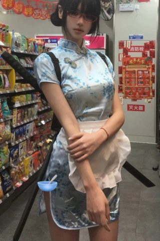 A doll wearing a light blue cheongsam with floral patterns, paired with a white apron, stands in a convenience store aisle.