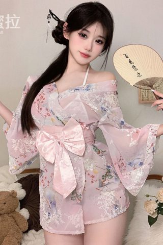 A woman poses in a light pink kimono-style pajama set with floral patterns, holding a fan and standing beside a teddy bear and flowers.