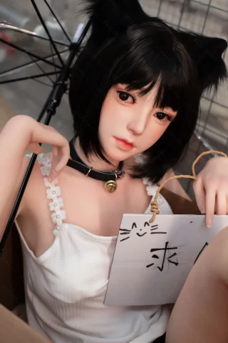 A doll with short black hair and cat ears sits in a box, holding a sign while wearing a white tank top and a choker.