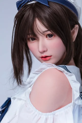 A doll with long, dark hair and soft facial features, wearing a white ruffled top with a blue accent, gazes over her shoulder.
