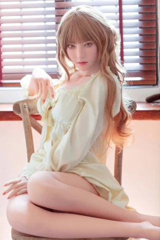 A doll with long, wavy blonde hair sits gracefully on a wooden chair, wearing a soft, pale yellow dress. She poses with one arm extended, fingers gently pointing, against a backdrop of wooden blinds and warm lighting.