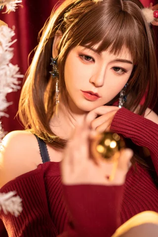 A young woman with brown hair and pigtails wears a deep red off-shoulder sweater, holding a golden ornament, with a festive background.
