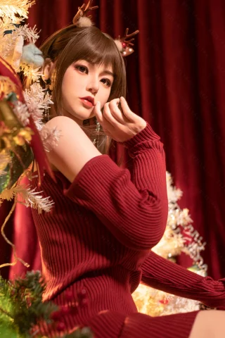 A doll with long brown hair and antler accessories poses in a cozy red sweater, sitting near a decorated Christmas tree with a rich red backdrop.