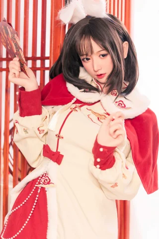A young woman poses in a festive outfit featuring a cream dress with red accents and intricate embroidery, complemented by a red cape with white fur trim. She holds a decorative card and has her hair styled in soft waves, adorned with a small headpiece resembling animal ears. The background features traditional wooden latticework, enhancing the celebratory atmosphere.