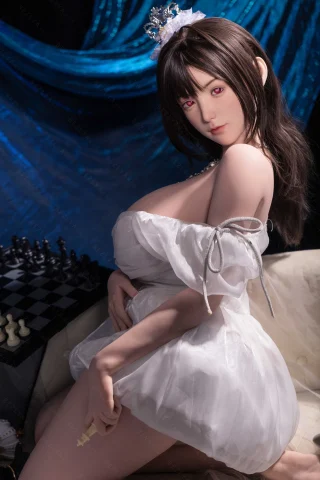 A doll with long dark hair and red eyes wears a white dress with puffed sleeves, sitting on a couch beside a chessboard.