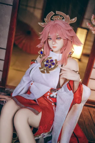 A doll with pink hair and ornate accessories sits gracefully, wearing a traditional outfit with red and white colors, in a serene indoor setting.