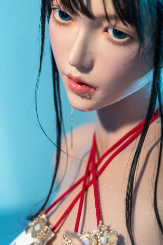 Praying amulet doll with long black hair, blue eyes, and red straps. Water droplets accentuate the skin, set against a blue backdrop.