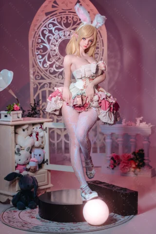 Doll with bunny ears in a frilly pastel dress, striking a playful pose on a round pedestal, surrounded by soft decor and plush toys.