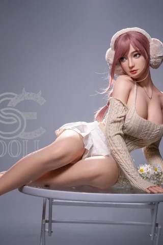 A lifelike doll with long pink hair, wearing a cozy off-shoulder sweater and a white skirt, poses gracefully on a round table. She has white earmuffs and is surrounded by soft lighting, highlighting her features.