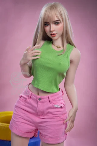 A doll with long blonde hair wears a green ribbed tank top and pink shorts, posing playfully against a pink background.