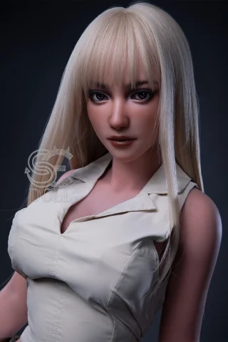 A lifelike doll with long, straight blonde hair and a subtle makeup look, wearing a fitted cream-colored blouse, against a dark background.