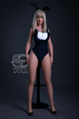 A doll with long blonde hair and a black bunny ear headband, wearing a fitted black swimsuit with a white ruffled collar, stands confidently beside a stool.