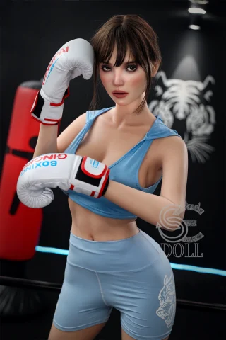A young woman in a light blue sports outfit poses with boxing gloves in a gym, featuring a boxing bag in the background.