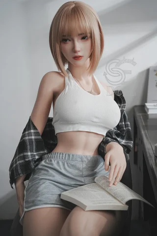 A doll with short blonde hair sits casually, wearing a white crop top and gray shorts, holding an open book, with a plaid shirt draped over her shoulders.