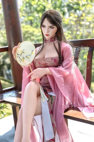 A doll with long dark hair, wearing a pink floral dress and matching robe, holds a decorative hand fan while seated outdoors.