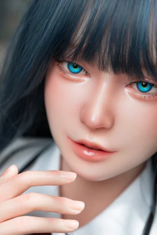 The image features a lifelike doll with long, dark blue hair and striking green eyes. The doll's skin has a smooth, realistic texture, and it is wearing a white shirt. One hand is gently touching its face, emphasizing a soft and serene expression. The background is softly blurred, focusing attention on the doll's features.