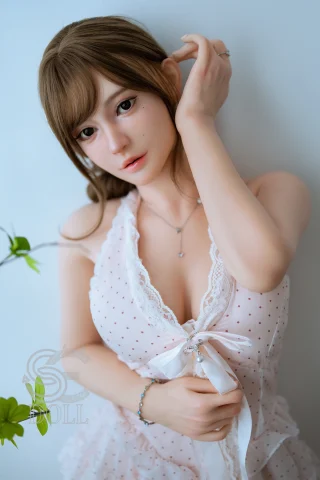 A doll with long brown hair, wearing a light pink polka-dot dress, poses with one hand near her face against a white background.