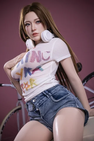 Miaoyu 171cm C-Cup doll wearing a white graphic t-shirt and denim shorts, seated with headphones, against a pink background.